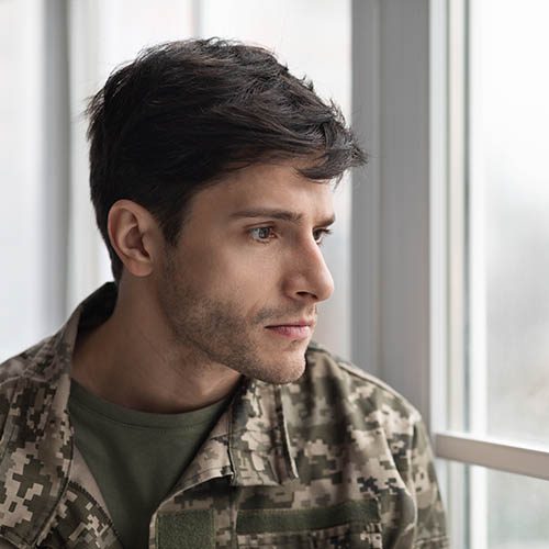 Unhappy young military man in camouflage uniform looking through window at home, feeling depressed after coming back from military service, panorama with copy space, closeup shot