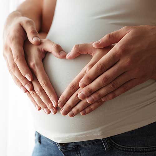 Close up portrait of man's and woman's hands making heart gesture over pregnant belly indoors