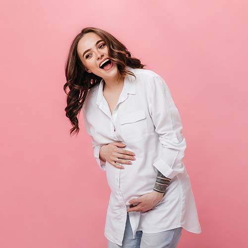 Portrait of emotional happy woman in white long shirt. Joyful pregnant lady in jeans laughing and posing on pink background