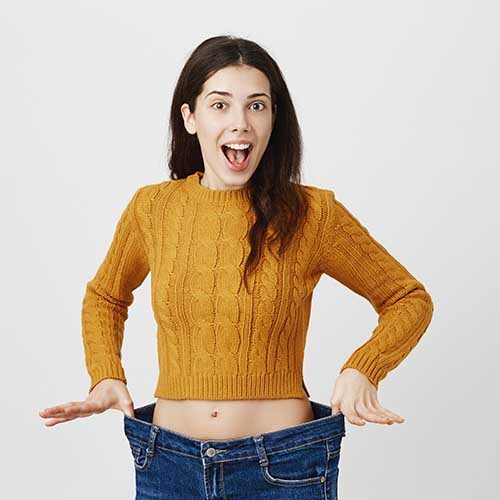 Astonished and happy young lady being impressed and excited because of losing weight, showing empty space in jeans by stretching it, standing over gray background. Girl is ready for summer.