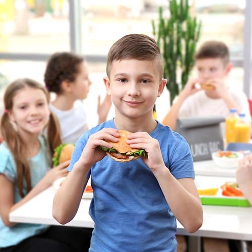 Cute boy eating hamburger and children sitting at table in school cafeteria